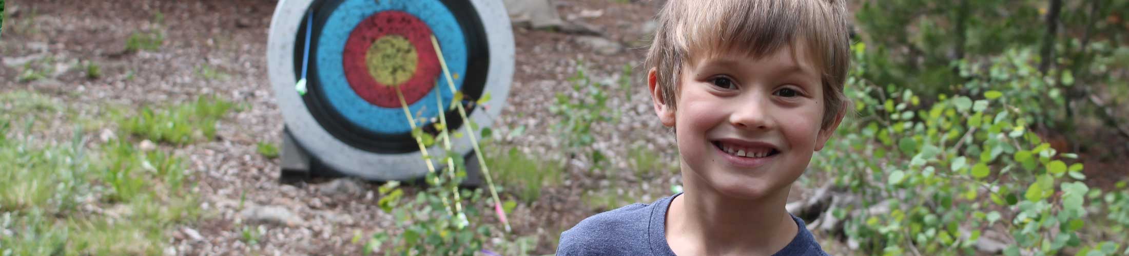 Young boy with colorful archer board with arrows in background at The Salvation Army High Peak Camp Colorado